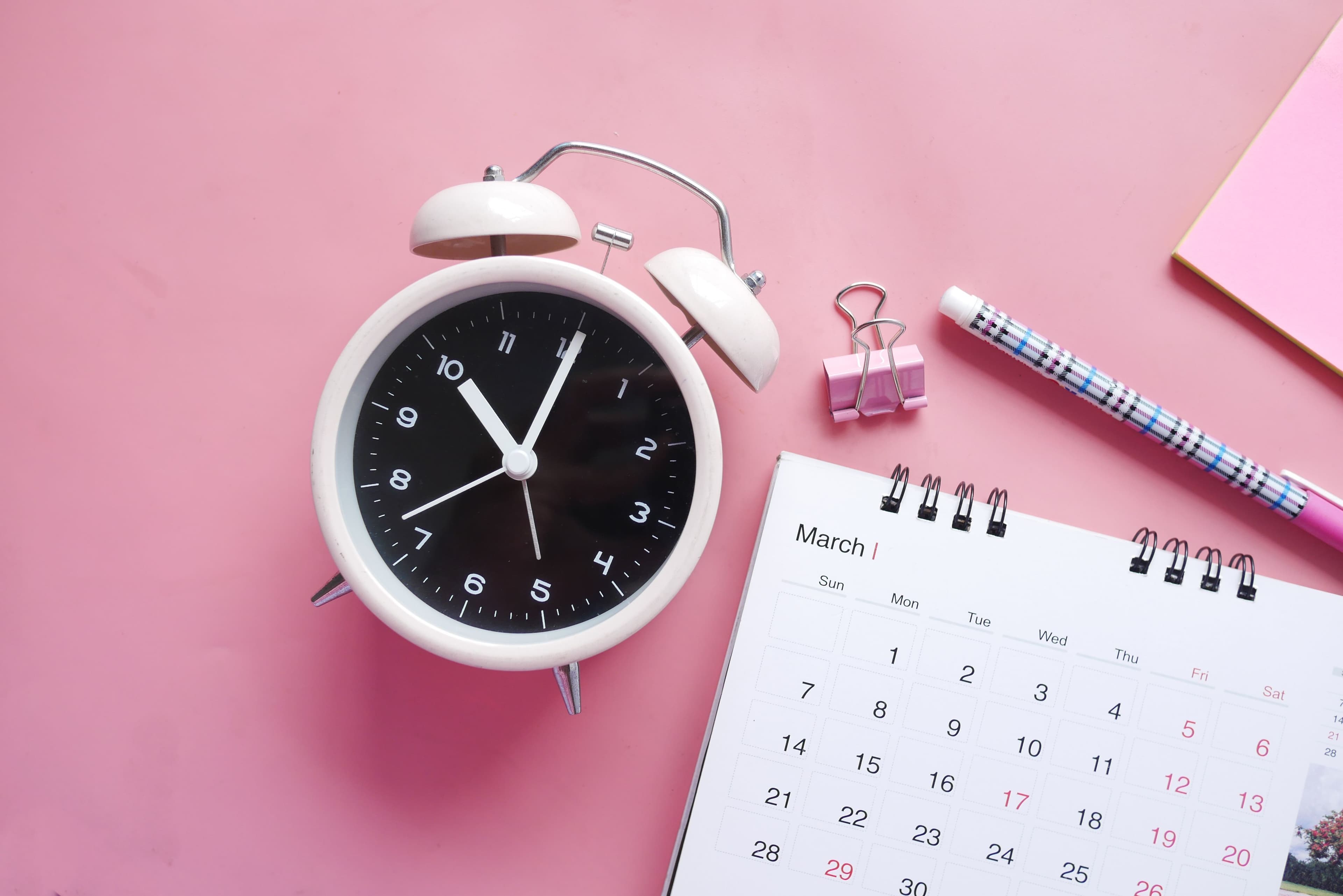 White alarm clock beside a March desk calendar, pink binder clip, and pen on a pink background.