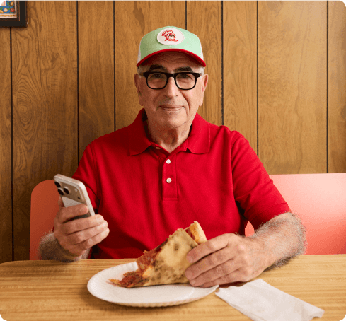 A pizzeria owner in a red polo shirt and green cap sits at a wooden table, holding his phone in one hand and a slice of pizza in the other.