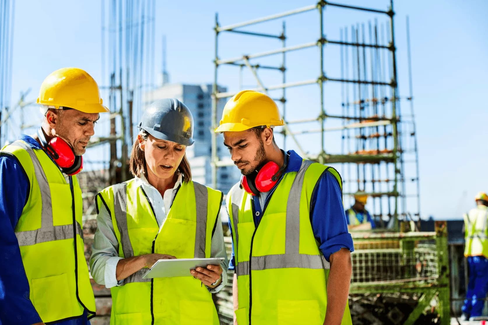 Three construction workers in safety vests and helmets reviewing a tablet at a construction site.