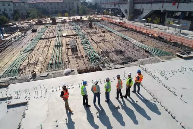Group of construction workers wearing safety gear inspecting a large building foundation with exposed rebar and piping.
