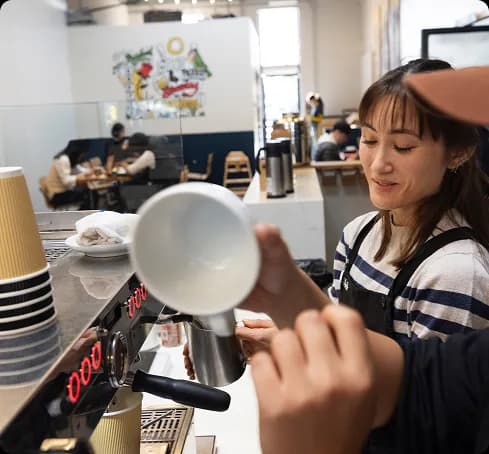 A barista in a striped shirt and apron holds a white cup at an espresso machine in a bright cafe with customers seated behind.