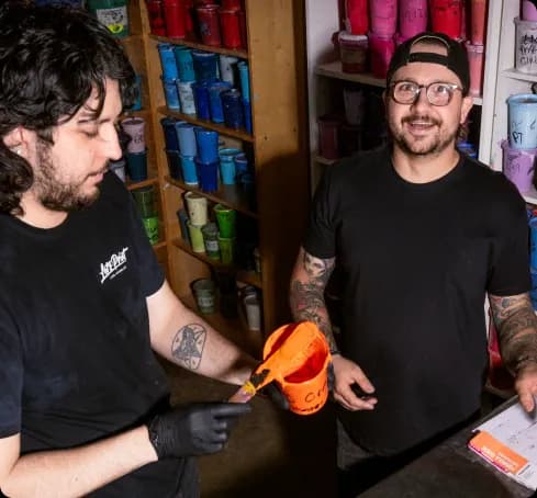 Two tattooed workers in black t-shirts collaborate in a paint or craft shop, one painting an orange bucket while the other looks on, with colorful cups lining the shelves behind them.
