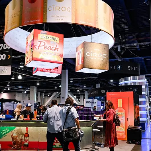 Trade show booth featuring illuminated hanging signs for Smirnoff Peach Lemonade and Ciroc vodka, with people engaging at the counter.
