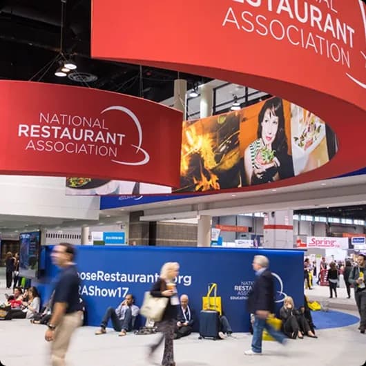 Convention scene with people walking and sitting near National Restaurant Association banners and booths.