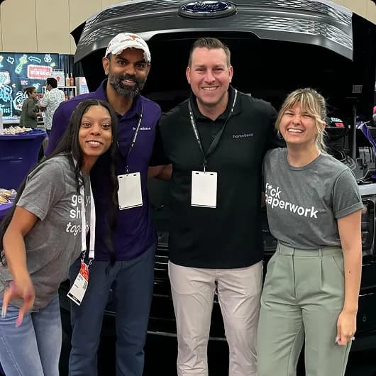 Four smiling people standing in front of an open Ford vehicle hood at an indoor event.