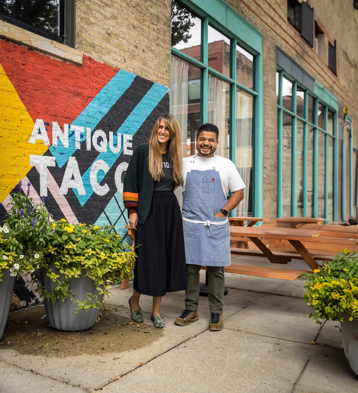 A smiling woman and man standing outside a restaurant with a colorful 'Antique Taco' mural on the brick wall.
