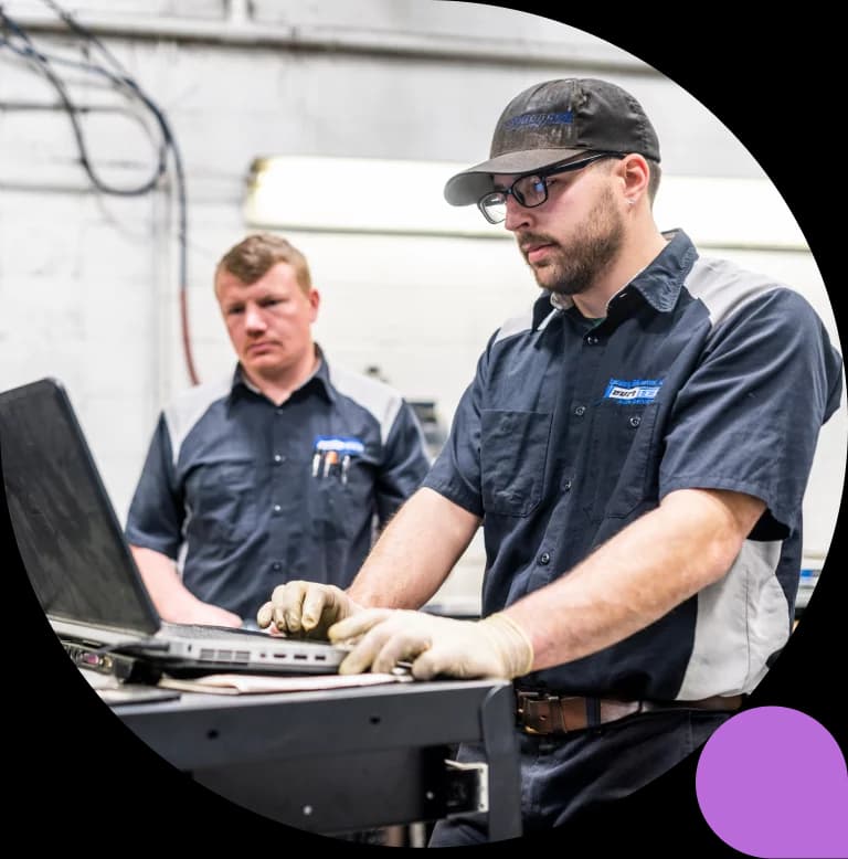 Two auto-shop technicians in work shirts at a bench, one entering details on a laptop while the other watches.