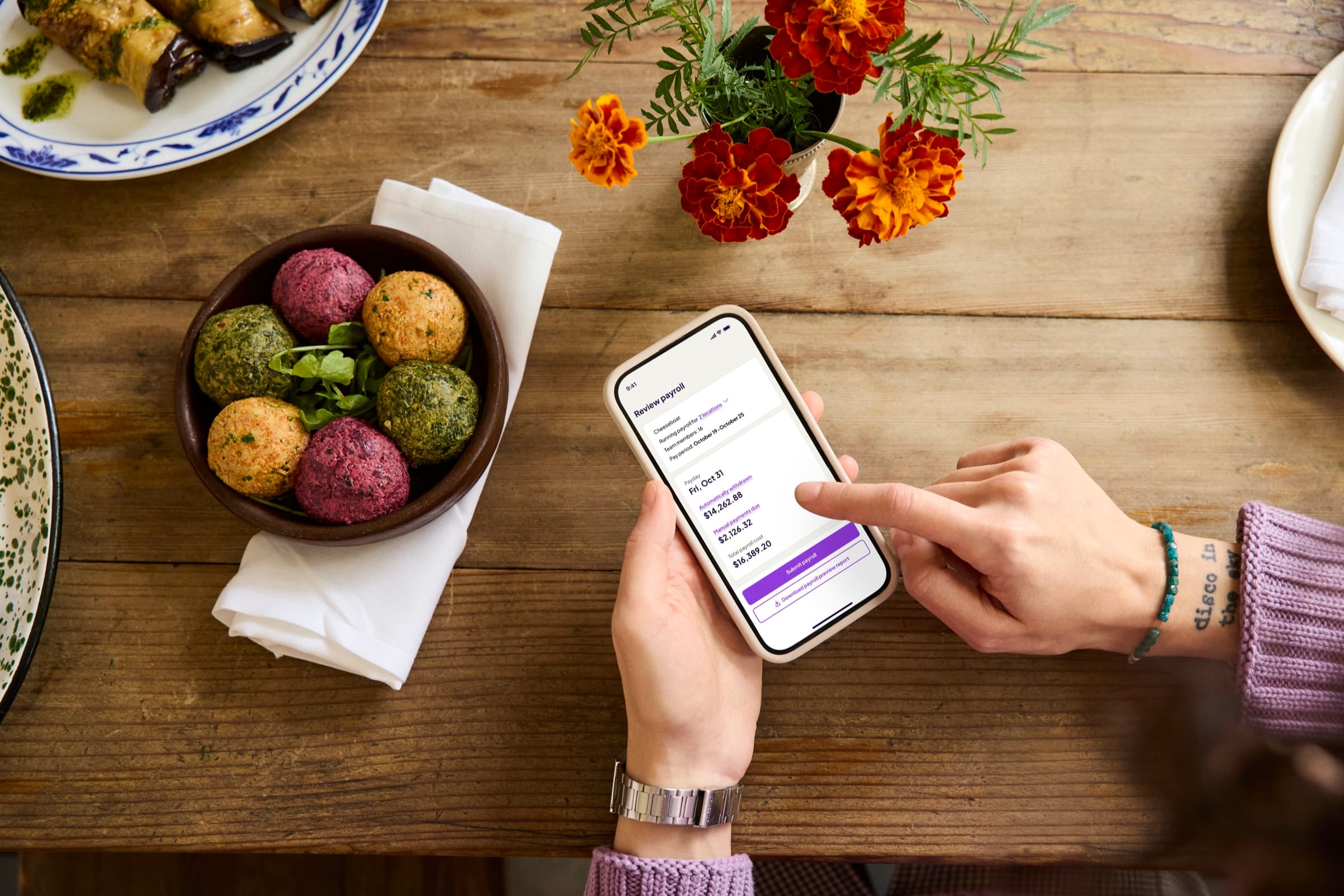 A person holding a phone showing Homebase payroll review next to a bowl of falafel on a restaurant table
