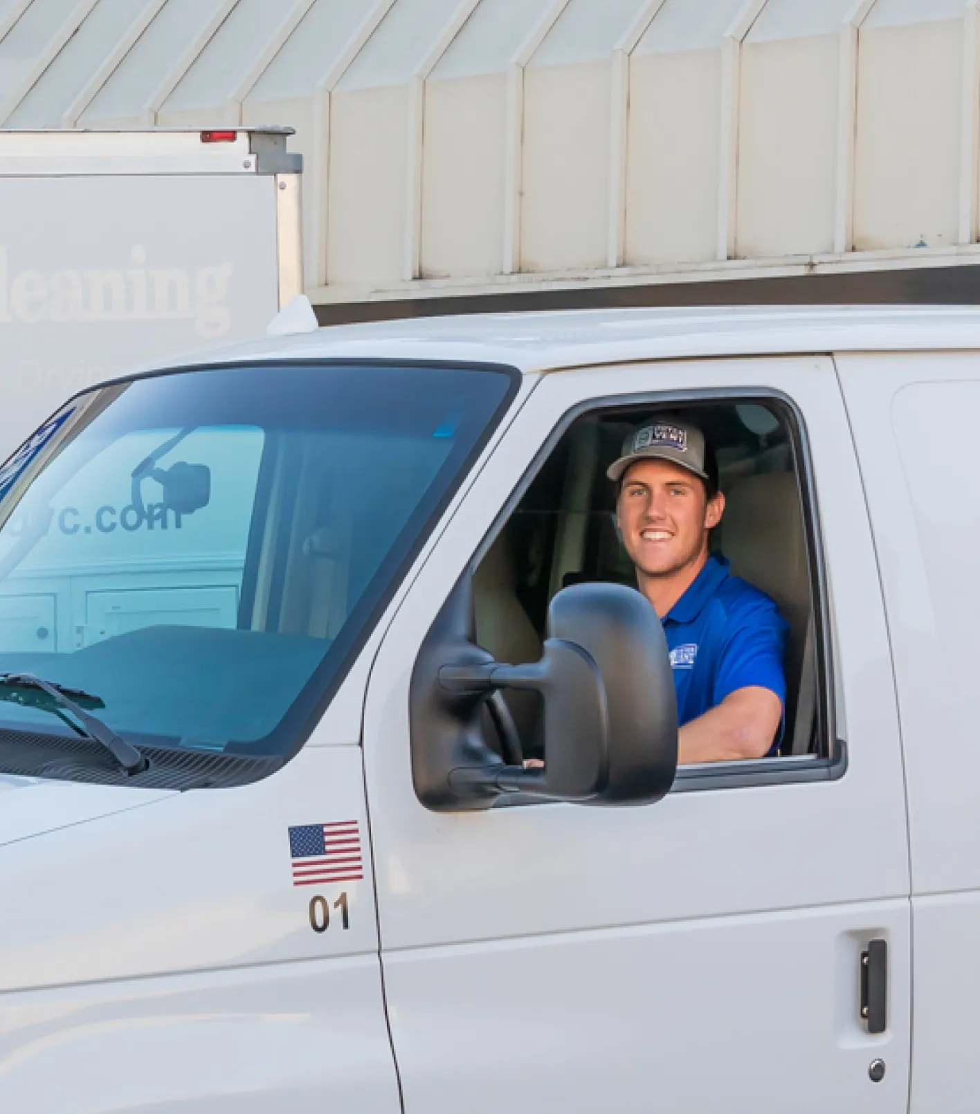 Smiling man in blue shirt and cap sitting inside a white service van with an American flag decal on the door.