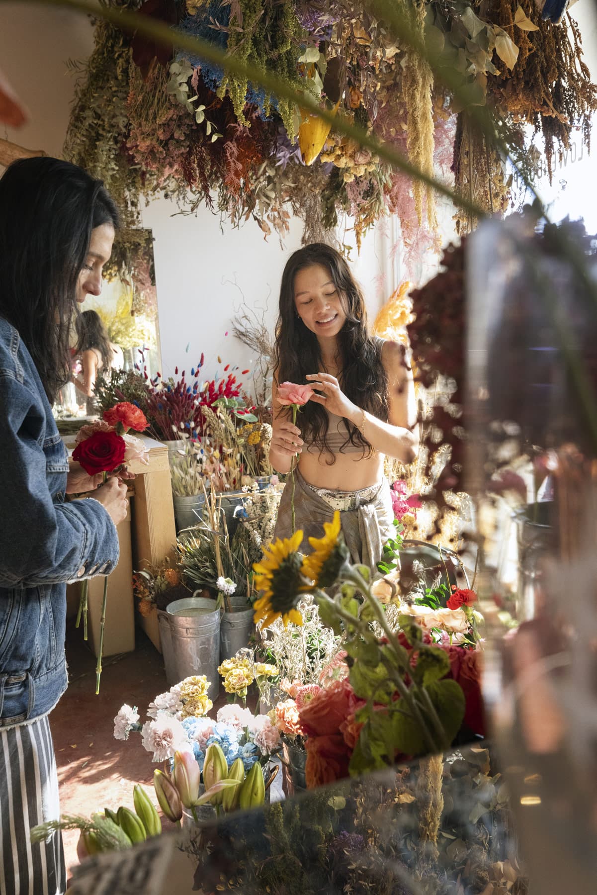 Two people arranging flowers in a florist shop filled with dried and fresh blooms, one holding a red rose and the other a pink flower.
