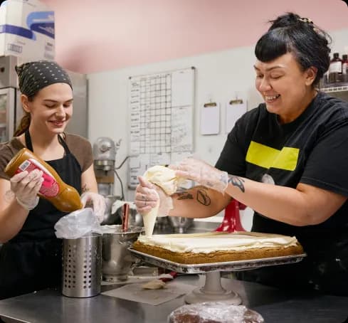 Two bakery workers in aprons laughing together as one pipes frosting over a large sheet cake in a commercial kitchen.