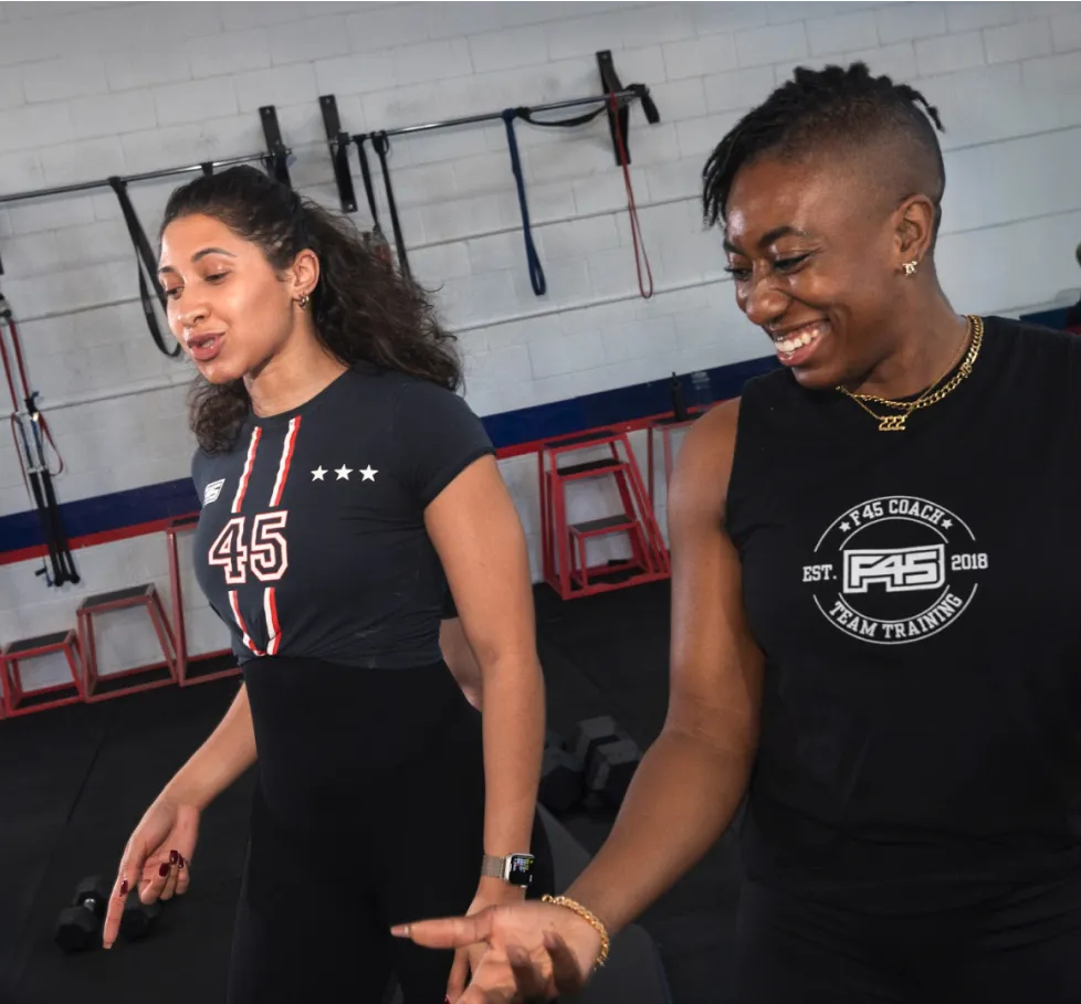 Two smiling women in workout gear at an F45 gym, one in a '45' jersey, standing near a wall of resistance bands and dumbbells.