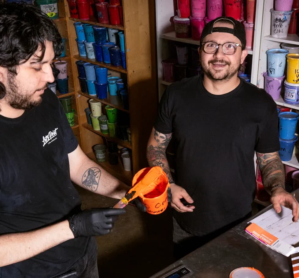 Two men laugh together at a print shop counter, one holding an open bucket of orange ink beside shelves stacked with paint tubs.