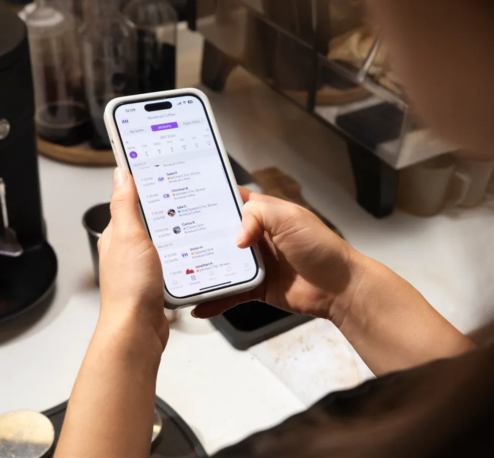 Hands hold a phone displaying a shift schedule app for Rosebud Coffee, listing team members' shift times, at a coffee shop counter.