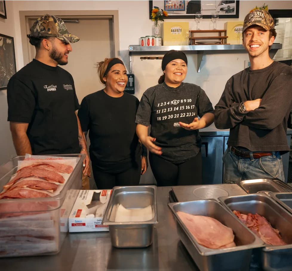 Four smiling deli or restaurant workers standing behind a stainless steel counter with trays of sliced meats.