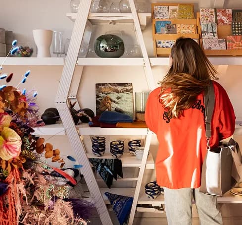 Shop worker in an orange jacket restocking shelves of ceramics, prints and dried flowers in a boutique.
