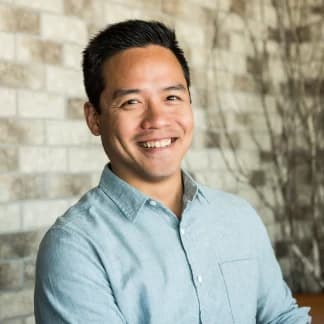 A smiling man in a light blue button-down shirt poses for a headshot in front of a brick wall.