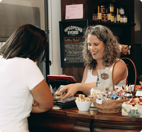A small-business owner smiles as she rings up a customer at her shop counter, a card reader and baskets of treats in front of her.