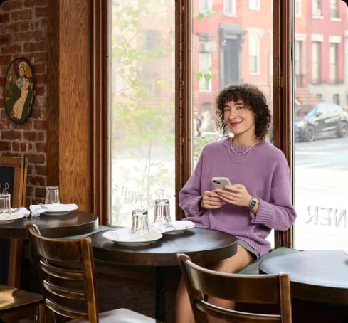 A woman in a purple sweater sits at a round table in a cozy cafe, smiling at her phone. Large windows reveal a street with red brick buildings.