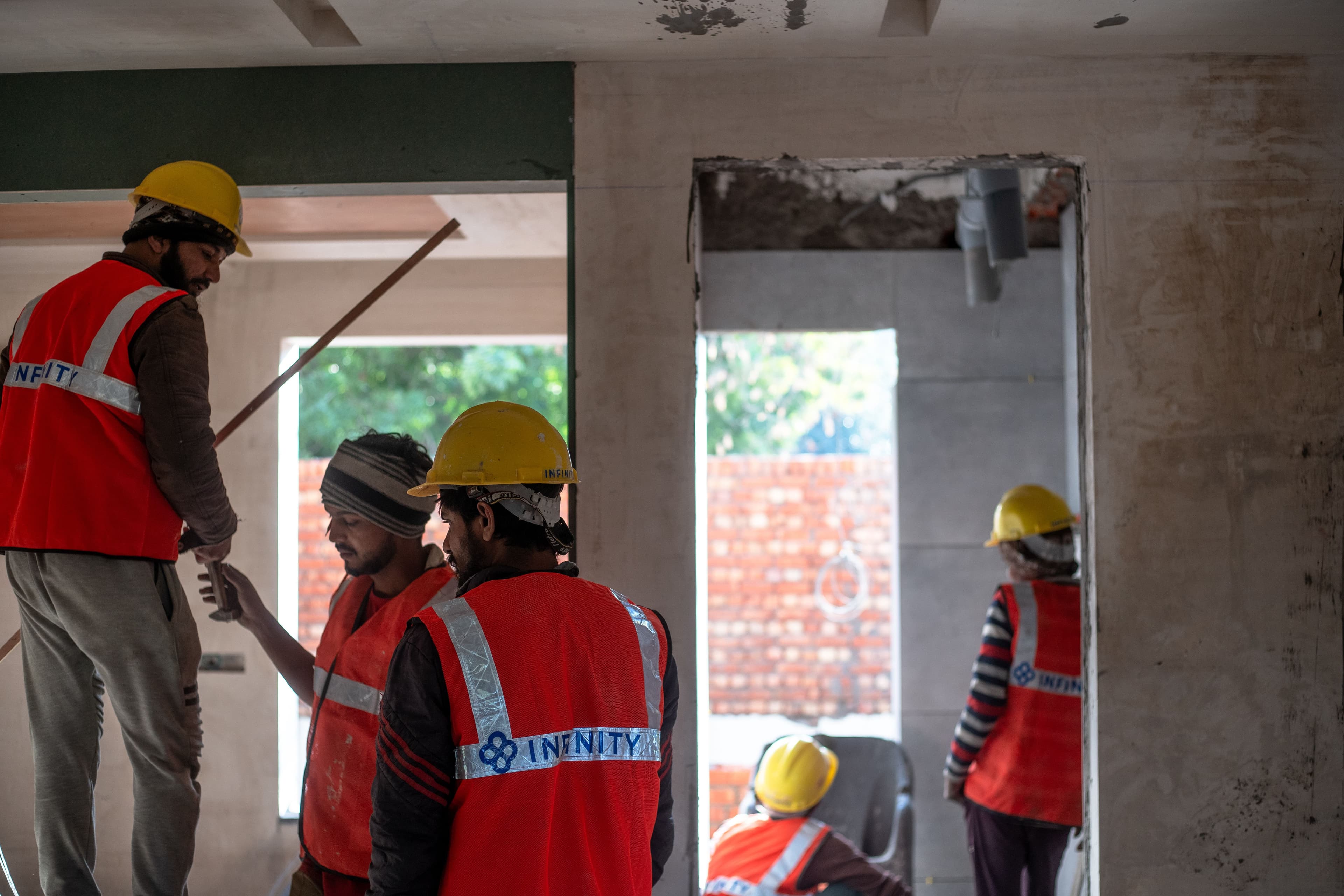 Construction workers in hard hats and orange safety vests gather inside an unfinished building.
