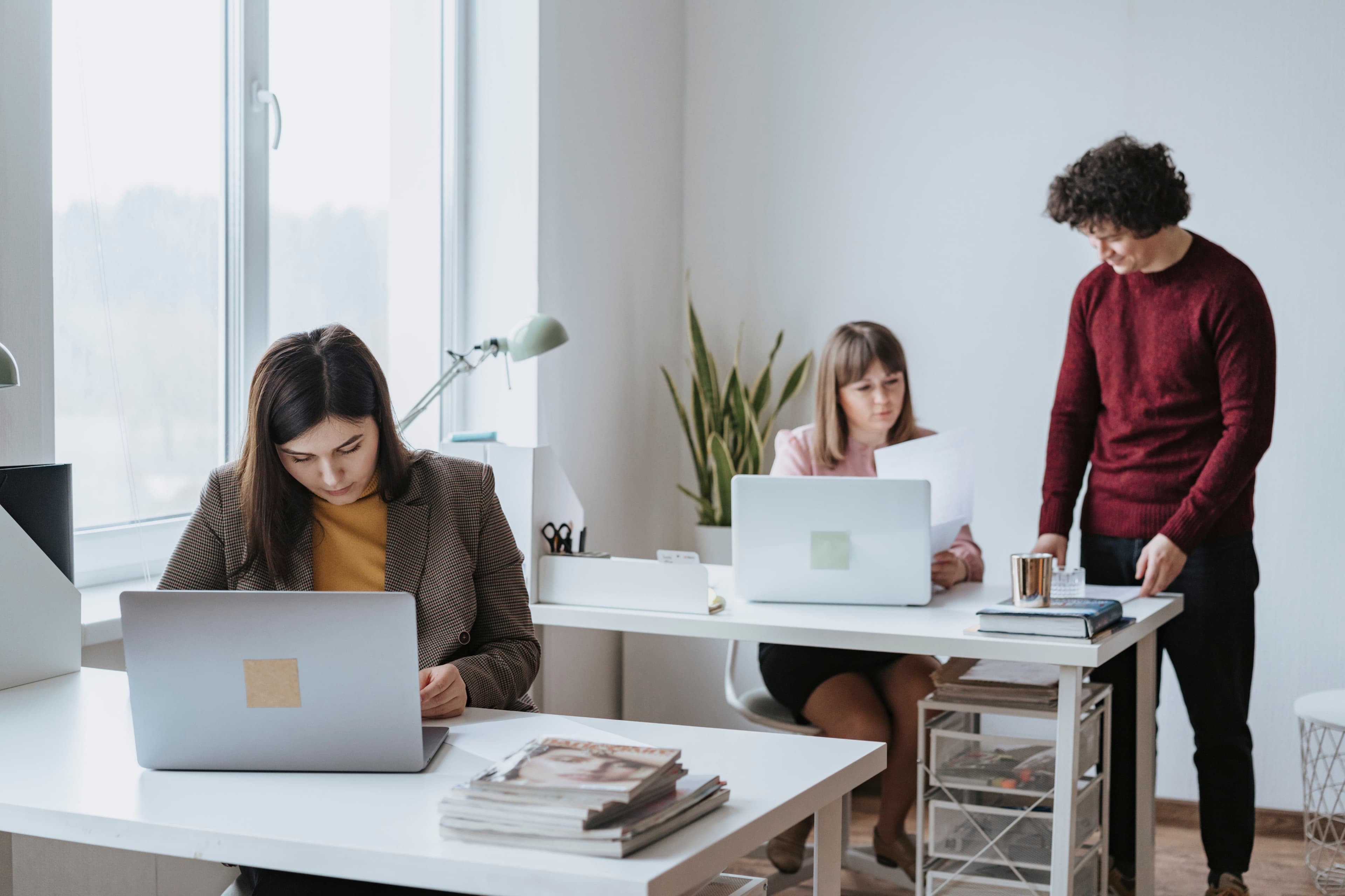 An image of three people in an office, one person working on their laptop on their desk, while two other people discussing a project at another desk.